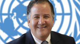 رجل مبتسم ينظر مباشرة إلى الكاميرا مع شعار الأمم المتحدة في الخلفية.