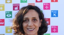 امرأة مبتسمة تقف أمام ملصق لأهداف الأمم المتحدة في مجال التنمية المستدامة.