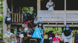 Les membres d'une famille, tous équipés d'un masque de protection respiratoire, sont assis sur la terrasse d'une maison en bois, tandis que plusieurs enfants portant également un masque marchent en direction de l'objectif en s'éloignent de la maison, divers objets distribués par l'UNICEF à la main.