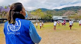 Au Costa Rica, une femme portant le gilet bleu de l’OIM et un masque chirurgical et faisant dos à la caméra regarde des enfants jouer au football sur un terrain en arrière-plan duquel on peut voir des collines verdoyantes.