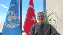 Official photo of the newly appointed Resident Coordinator for Turkey, Alvaro Rodriguez. He sits at a desk hands folded smiling in front the UN and Turkey flags.