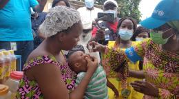 Un niño pequeño recibe la vacuna contra la polio mediante gotas en la boca, mientras su madre lo sostiene.
