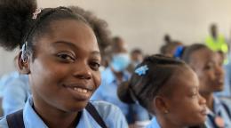 Gros plan sur une jeune fille haïtienne en uniforme scolaire souriant à la caméra tandis que d'autres élèves, en arrière-plan, regardent en direction de l'enseignant.