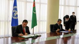 Deux hommes, des personnalités officielles, signent des documents dans un bureau, avec, derrière eux, le drapeau de l'ONU et celui du Turkménistan. 