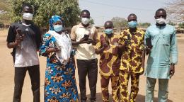 In Benin, a group of people wearing facial masks are standing outside, holding cell phones and looking at the camera.