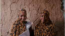Two girls smile as they sit on the floor against a wall as they hold documents. 