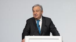 A man in a suit speaks into a microphone and a podium with UN logos on it, against a grey wall. 