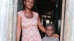 Una joven liberiana está de pie en la puerta de su casa, con el brazo alrededor del hombro de su hijo pequeño.