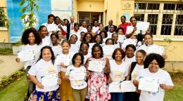 Women in white t-shirts hold certificates in their hands, in front of a municipal building, with the RC standing in the center-back.