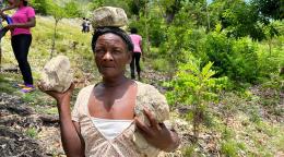 Une femme haïtienne se tient face à la caméra, au milieu d'un terrain escarpé jonché d'arbustes, en tenant une grosse pierre dans chaque main.