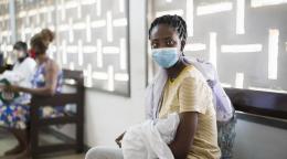 A woman wearing a respiratory protection mask and carrying her baby in her arms sits in a medical waiting room with other women.