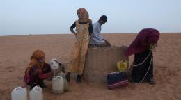 En un desierto de arena roja, cuatro jóvenes intentan llenar jarras de agua de un pozo.