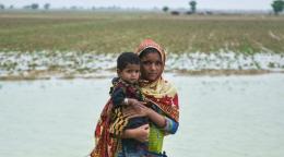 A girl in scarf holds a toddler in her arms in front of a pool of water collected on a field, against a grey sky. 
