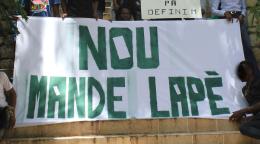 Un plano detalle de una gran pancarta con grandes letras verdes, que es sostenida por activistas juveniles quienes exigen paz a través de un eslogan escrito en criollo.