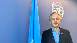 Un Coordinador Residente-funcionario de las Naciones Unidas se encuentra frente a una pared azul y una bandera azul de las Naciones Unidas.