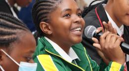 Молодая намибийская девушка в зеленом спортивном костюме с улыбкой говорит в микрофон.