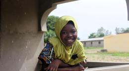 Une jeune fille portant un voile de couleur claire autour de la tête est penchée au-dessus d'un balcon dans une campagne togolaise et regarde en direction de la caméra en souriant.