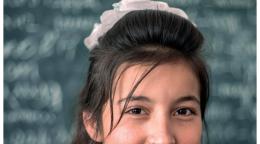 Portrait d'une jeune fille brune aux cheveux attachés, souriant à la caméra devant un tableau de classe.