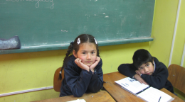 Una niña y un niño sentados en sus pupitres frente a un pizarrón en un salón de clases.
