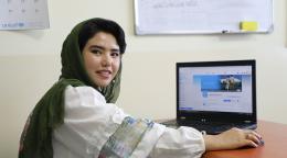 Una joven afgana sonríe a la cámara, con un ordenador portátil sobre el escritorio al fondo.