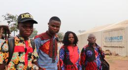 In Angola, two young Congolese men stand with their mother and father outside a UNHCR refugee camp. The four family members wear brightly colored clothes and look at the camera.