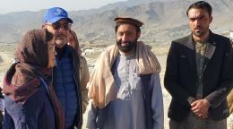 Dans un vallée, au pied d'une chaîne montagneuse, une femme portant un foulard discute avec trois hommes, l'un vêtu d'une veste bleue et d'un casquete et les deux autres de tenues traditionnelles afghanes.