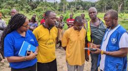 In Liberia, on the edge of a forest, two men wearing yellow t-shirts and a female UN Women staff member watch a third man, a WFP staff member, write in a notebook, while a group of people stand in the background. 