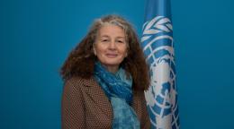 Una mujer con chaqueta marrón y pañuelo azul junto a una bandera de las Naciones Unidas.