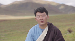 Un homme tibétain est assis à même le sol, au milieu d'une plaine, non loin d'une montagne que l'on peut apercevoir en arrière-plan. 