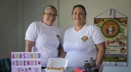 Two women in white shirts standing in a bakery kitchen with kitchen tools and baked goods