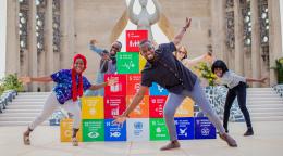 Four children playfully stand in front of a series of block with the SDGs
