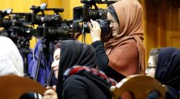 Woman in a brown dress and headscarf peering beneath a journalist's camera in Afghanistan