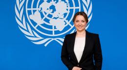 Woman in a black suit stands against a blue background with the UN logo
