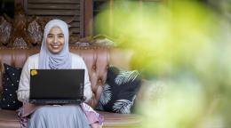 Woman in a white and grey headscarf sits on a bench with a laptop in her lap