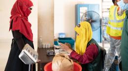 A woman in a yellow headscarf checks another woman in a red scarf's papers at a center for food items.