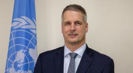 A UN official in a suit and tie poses for a photo next to a blue UN flag