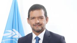 Man in a dark blue suit and blue tie standing in front of a light blue UN flag