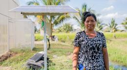 A woman in a black printed dress stands in front of a solar panel installed in a green field in rural Cambodia