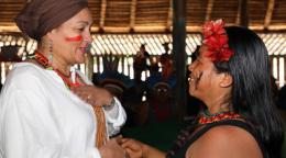 A woman in a white shirt and red headscarf shakes hands with a shorter woman in red headfeathers and face markings