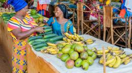 Two women in colourful clothes laugh with each other in a market place, behind a stall selling mangoes and bananas