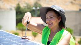A woman in a hat and a green vest works with a tool fixing a solar panel