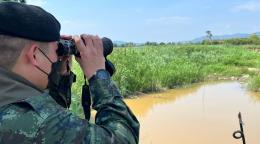 A man wearing a mask, dressed in camo-military garb and a black beret, looks out at a murky river with binoculars.