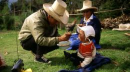 A man in a hat feeds an infant while a woman sits by the infant on a grass area in the sun.