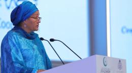 A woman in a blue cover-up and glasses and dangling earrings speaks at a podium.