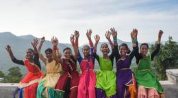 group of women wearing colourful saris smile to the camera with their hands in the air.