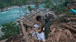 A woman in a green dress kneels over some destroyed wood right by a flowing blue river in Nepal.