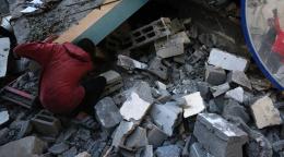 A young boy in a red shirt kneels among the rubble, stones and broken building, searching for something.
