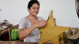 A woman in a white shirt weaves together a yellow star from reeds.