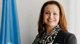 A woman with brown hair and a black suit and scarf, the UN Resident Coordinator in UAE, stands next to a blue UN flag.
