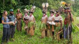 A group of indigenous peoples in traditional clothes and headgear, speak to a man and a woman dressed in dark blue attire. They are all standing in a lush green forest clearing.
